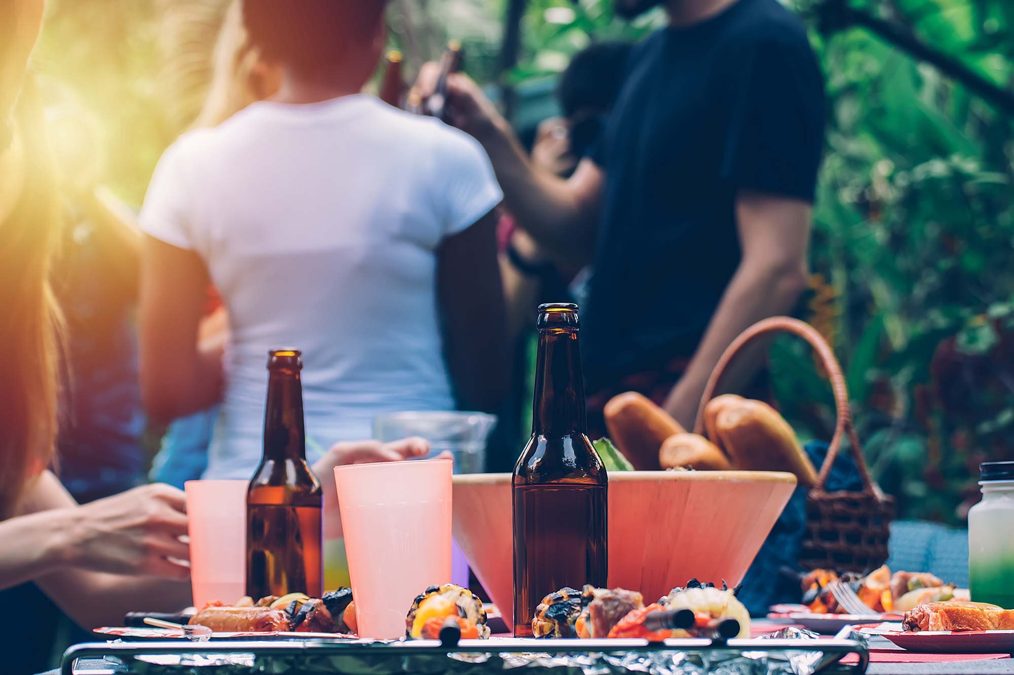 Kanutour und Planwagenfahrt mit Grillplatz Grillabend im Freien mit gegrilltem Gemüse, Bierflaschen und Menschen, die im Hintergrund unter Bäumen stehen.
