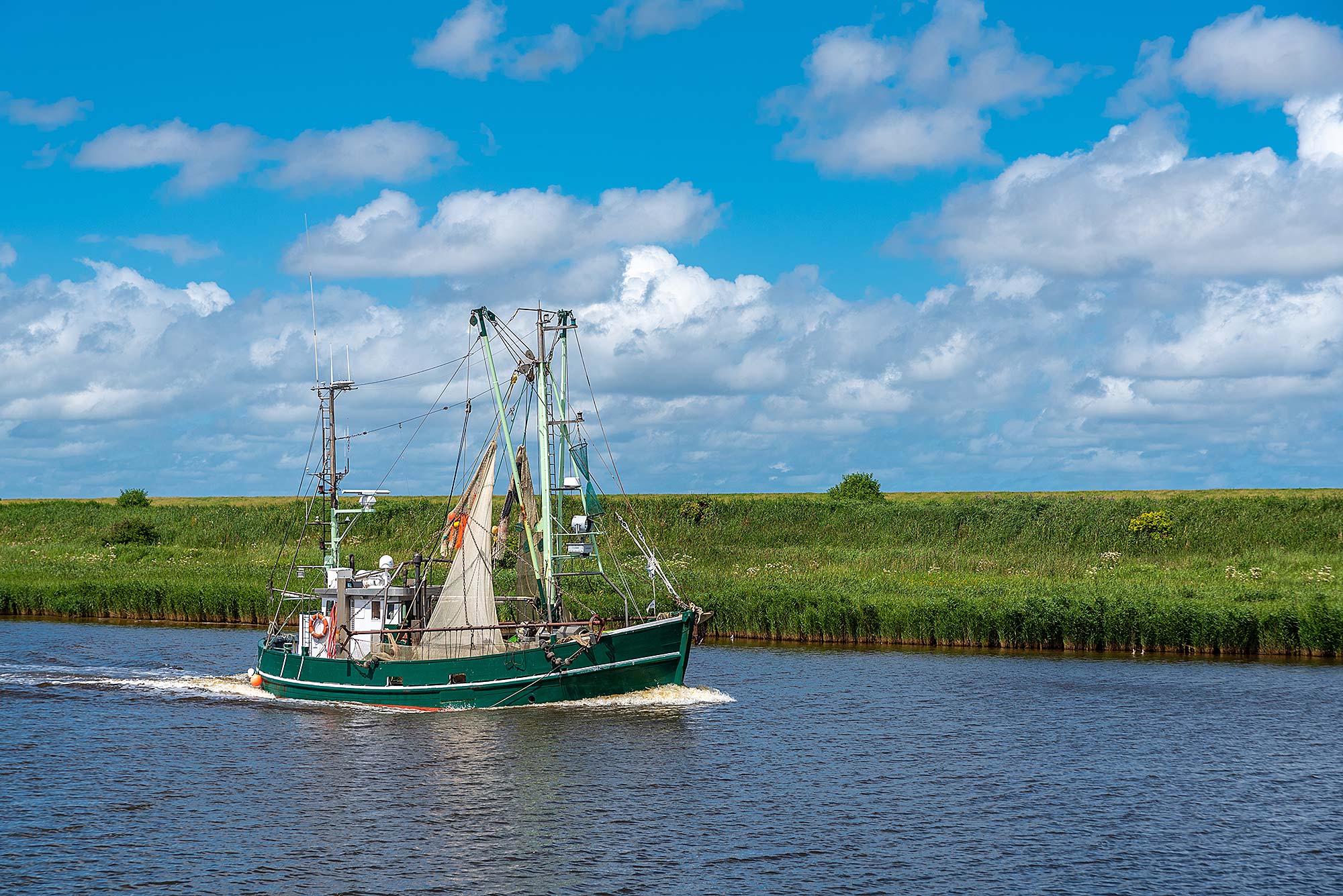 Kanutour und Kutterfahrt Grünes Fischkutter fährt auf einem Fluss unter blauem Himmel mit weißen Wolken, im Hintergrund eine grüne Wiese.